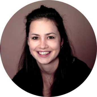 Porträt einer Person mit dunklem Haar, die freundlich lächelt. Der Hintergrund ist dunkel und leicht unscharf.