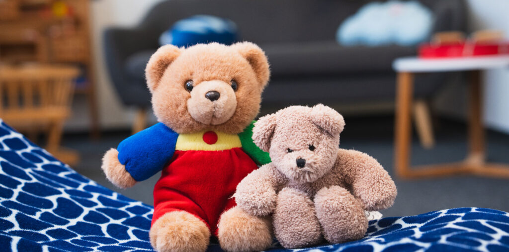 Zwei Teddybären sitzen nebeneinander auf einer Decke, im Hintergrund ein Spielraum.