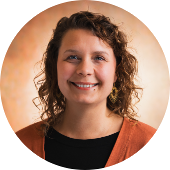 Porträt einer Person mit lockigem Haar, die lächelt und eine dunkle Kleidung mit orangefarbener Strickjacke trägt. Der Hintergrund ist weich und hell.