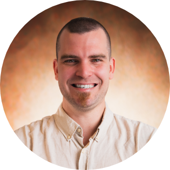 Porträt einer Person mit sehr kurzen Haaren und hellem Hemd, die freundlich in die Kamera lächelt. Der Hintergrund ist warm und neutral gehalten.