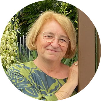 Porträt einer älteren Person mit blond-grauem Haar und Brille, die lächelnd an einem Holzbalken steht. Im Hintergrund sind grüne Pflanzen zu sehen.